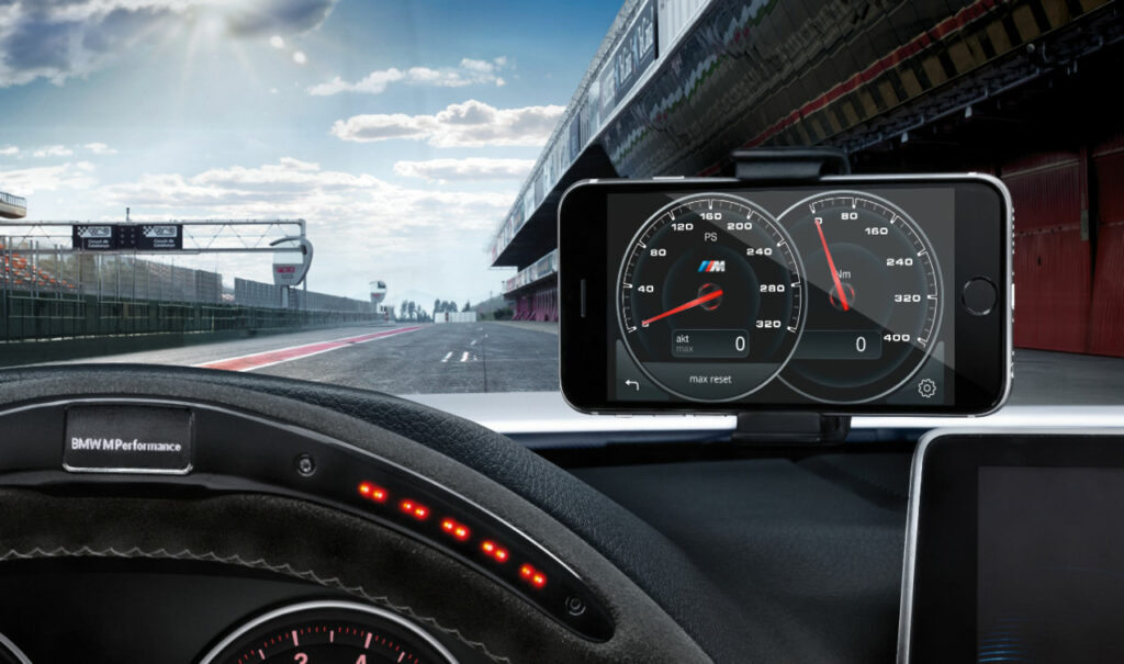 BMW M Performance Drive Analyser 8 Ein auf dem Armaturenbrett montiertes Smartphone zeigt mithilfe des BMW M Performance Drive Analysers Geschwindigkeits- und Drehzahlwerte an. Durch die Windschutzscheibe bei leicht bewölktem Himmel ist eine Rennstrecke sichtbar. Auf dem Lenkrad ist BMW M Performance zu sehen.