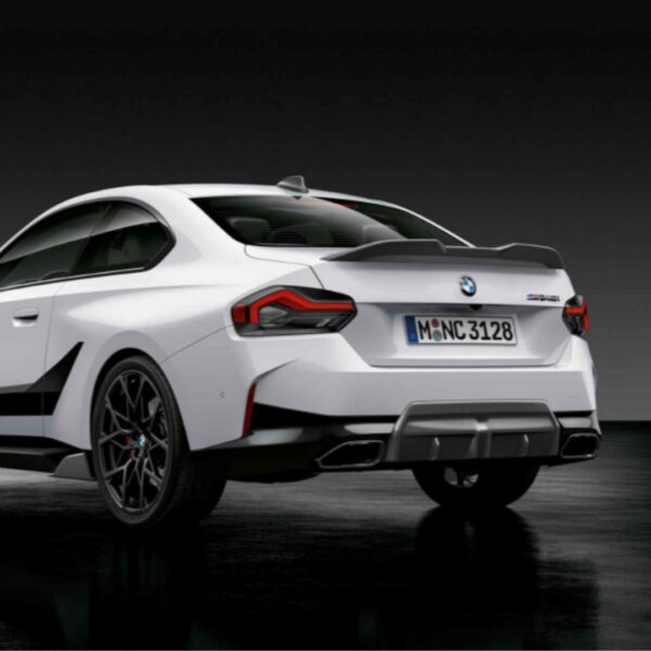 Von hinten links ist ein weißer Sportwagen BMW M2 zu sehen, der sein sportliches Design, die Doppelauspuffrohre, den Heckspoiler und das deutsche Nummernschild mit der Aufschrift M Ø NC 3128 vor einem dunklen Hintergrund zur Schau stellt.