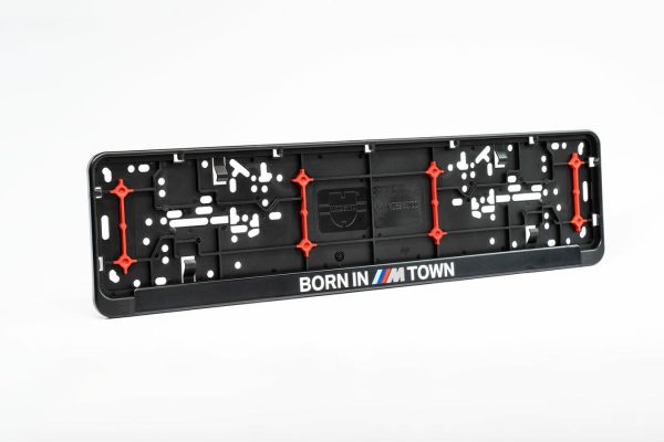 Der Kennzeichenhalter born in m town 2 ist ein schwarzer BMW-Kennzeichenhalter mit roten Akzenten und dem Schriftzug "BORN IN ///M TOWN" in weißer, blauer, lila und roter Schrift am unteren Rand, dargestellt auf einem schlichten weißen Hintergrund.