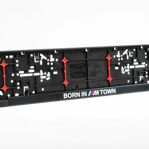 Der Kennzeichenhalter born in m town 2 ist ein schwarzer BMW-Kennzeichenhalter mit roten Akzenten und dem Schriftzug "BORN IN ///M TOWN" in weißer, blauer, lila und roter Schrift am unteren Rand, dargestellt auf einem schlichten weißen Hintergrund.